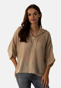 Pull à capuche en tricot beige avec fermeture éclair, design surdimensionné et manches courtes. Présente une finition texturée et un accent logo subtil.