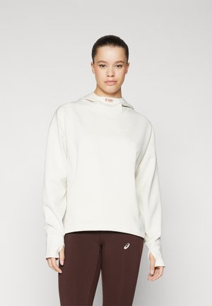 Cream hoodie with a high neckline, subtle logo, and long sleeves, paired with brown leggings featuring a small logo on the ankle.