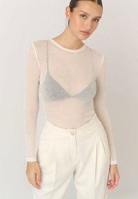Przezroczysta bluzka z długim rękawem w jasnym beżowym kolorze z okrągłym dekoltem, w połączeniu z białymi spodniami o wysokim stanie z pojedynczym guzikiem i fałdami.
