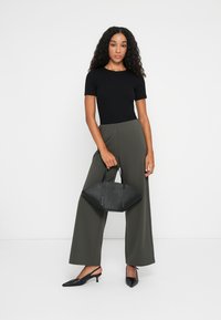 Schwarzes, figurbetontes Kurzarmshirt, hochtaillierte, olivfarbene weite Hose, schwarze spitze Ballerinas, hält eine strukturierte schwarze Handtasche mit kurzen Griffen.