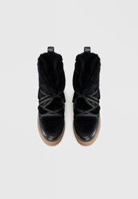 Une paire de bottes d'hiver en fourrure noire, avec des semelles épaisses couleur tan et des lacets noirs, montrée de dessus sur un fond blanc.