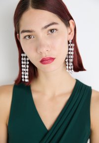 Rochie din material verde cu un decolteu în V. Subiectul are părul lung și drept de culoare marsala și poartă cercei lungi cu cristale argintii.