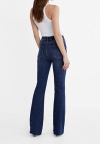 Jean flare en denim foncé à taille haute, avec des poches arrière et une finition épurée. Le tissu a une texture lisse et un léger stretch.