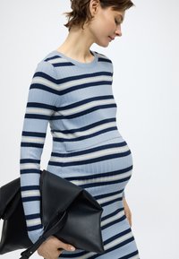 Ριγέ φόρεμα εγκυμοσύνης σε ανοιχτό μπλε, ναυτικό μπλε και κρεμ. Ύφασμα με ραβδώσεις και μακριά μανίκια. Το μοντέλο κρατά μια μαύρη δερμάτινη τσάντα.