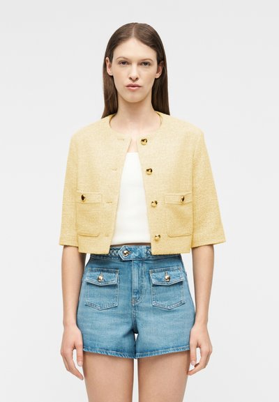 Femme portant une veste courte texturée jaune avec des boutons dorés sur un haut blanc, associée à un short en denim bleu taille haute avec des poches à boutons.