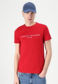 Sarkana kokvilnas t-krekls ar balto uzrakstu "TOMMY HILFIGER EST. 1985". Saskaņota ar zilu cepuri, kurai ir neliela logotipa detaļa.