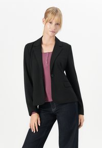 Kvinne med blondt hår iført en svart blazer over en lilla skjorte med knapper og mørke jeans, stående mot en enkel hvit bakgrunn.
