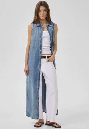 Kobieta ubrana w bezrękawnik z długiego dżinsu na białym topie na ramiączkach, białe szerokie spodnie, czarny pasek i czarne klapki, stojąca na jednolitym tle.