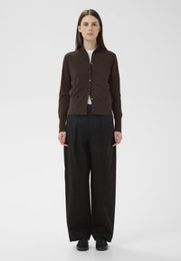 Barna kardigán gombos elejű és galléros, bő fekete nadrággal és fekete cipővel párosítva. Sima textúrájú, deréknál testhezálló.