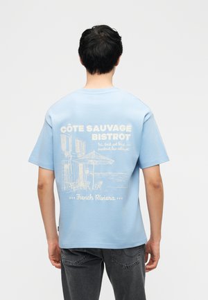 Άνδρας που φοράει ανοιχτό μπλε μπλουζάκι με άσπρο ναυτικό σχέδιο bistro, στέκεται γυρισμένος με την πλάτη, μπροστά σε απλό ανοιχτό γκρι φόντο.