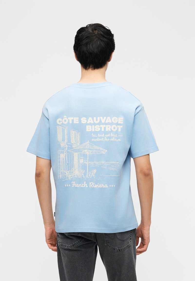 Άνδρας που φοράει ανοιχτό μπλε μπλουζάκι με άσπρο ναυτικό σχέδιο bistro, στέκεται γυρισμένος με την πλάτη, μπροστά σε απλό ανοιχτό γκρι φόντο.