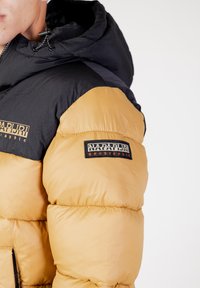 Giacca a vento con cappuccio e maniche nere, corpo color beige dorato, texture trapuntata e patch del logo sulla spalla sinistra.