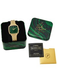 Montre carrée dorée avec un cadran vert à motifs et un bracelet en maille, présentée dans une boîte en métal marbrée verte et noire avec un certificat et une carte.