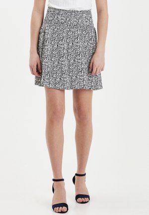 Femme portant une mini-jupe fleurie noire et blanche, un haut blanc et des sandales noires à bride à la cheville, debout devant un fond uni.