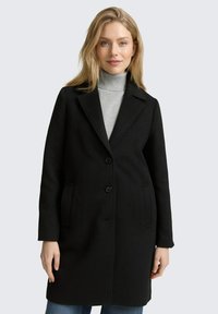 Cappotto di lana nera con colletto a punta, due tasche frontali e tre bottoni. Indossato sopra un dolcevita grigio, esposto su uno sfondo neutro.