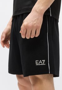 Čierne športové šortky s voľným strihom, bielym bočným pásikom a logom EA7 Emporio Armani na spodnej ľavej strane. Vyrobené z ľahkého materiálu.