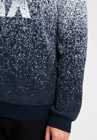 Marinblå och vit prickig stickad tröja med ribbad mudd, buren av en person med synlig hand och svarta byxor.