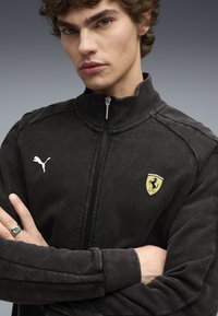 Sort zip-up jakke med høj krave, der har et hvidt Puma-logo og et gult Ferrari-badge på venstre bryst. Blød tekstur, sporty design.