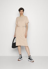 Vestido t-shirt bege até ao joelho com laço na cintura, mangas curtas e textura suave. Combinado com ténis pretos de cano alto e uma mala preta.