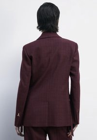 Blazer scozzese bordeaux con vestibilità su misura. Presenta un colletto a punta, due ventagli posteriori e bottoni dorati decorativi sui polsini.