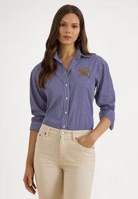 Modrá a biela prúžkovaná košeľa na gombíky s golierom a zlatým vyšívaným detailom. Kombinovaná s ľahkými béžovými nohavicami, ktoré majú uvoľnený strih.