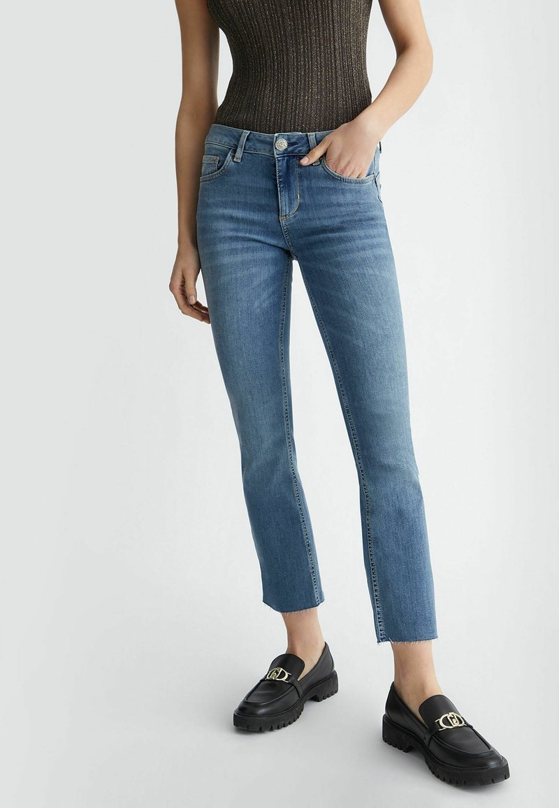 Högmidjade blå jeans med rak passform, fransad nederkant och två framfickor. Bärs med svarta loafers med en guldig accent.