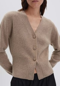 Femme portant un cardigan en tricot côtelé beige avec trois boutons, associé à un pantalon noir, sur un fond clair uni.