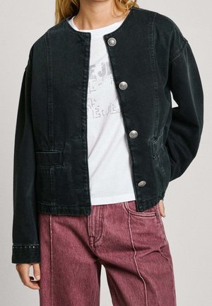 Personne portant une veste en denim noire boutonnée par-dessus un t-shirt blanc à motif, associée à un jean rouge délavé taille haute, debout avec une main dans la poche.