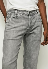 Jeans de mezclilla gris con un acabado ligeramente desgastado, que cuentan con cinco bolsillos, cierre de botón metálico y costuras visibles a lo largo de las uniones.