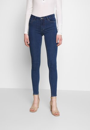 Vrouwelijk model draagt donkerblauwe skinny jeans gecombineerd met nude hoge hak sandaal en een wit shirt met lange mouwen, staand tegen een witte achtergrond.