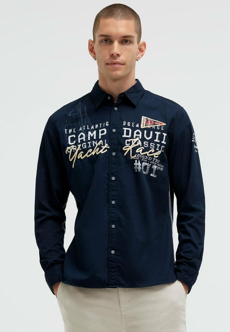 Marineblaue Hemdbluse mit Knopfleiste, bestickt mit Text und nautischen Motiven. Baumwollstoff, lange Ärmel, Standardkragen und tailliertes Design.