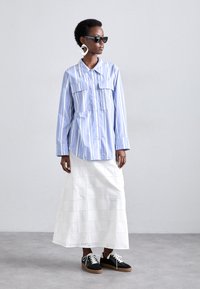 Blauw-wit gestreept overhemd met knopen en twee borstzakken, gecombineerd met een witte oogjes midi rok en zwarte sneakers met witte veters.