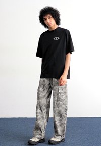 Black short-sleeve t-shirt with a graphic logo, paired with light grey camo pants featuring cargo pockets and a relaxed fit, worn with black shoes.