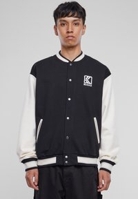 Veste varsity noire et crème avec fermeture à boutons, poignets côtelés et poches latérales. Présente le logo KANI sur la poitrine gauche. Texture de tissu douce.