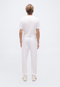 Άτομο που στέκεται με την πλάτη γυρισμένη, φορώντας λευκή καθημερινή στολή με κοντομάνικο πουκάμισο, χαλαρό παντελόνι και λευκά αθλητικά παπούτσια σε απλό φόντο.