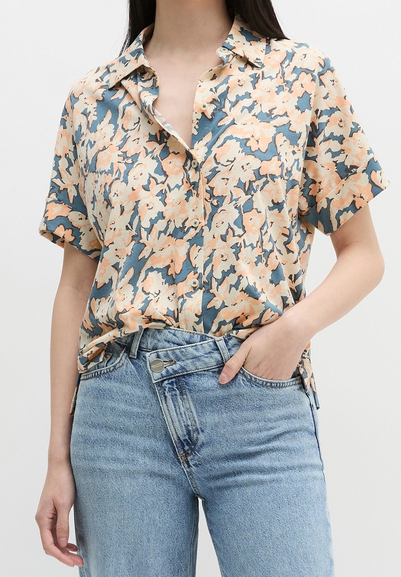 Femme portant une chemise fleurie à manches courtes boutonnée, aux couleurs pêche et bleu, rentrée dans un jean taille haute bleu clair.