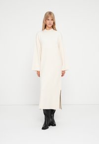 Robe longue crème avec un col haut, des manches trois-quarts et des fentes sur les côtés, associée à des bottes noires jusqu'aux genoux pour un contraste.