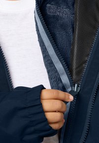 Giacca zip navy con fodera in morbido pile, caratterizzata da una texture a contrasto e una mano che tiene il tirantino della cerniera. Camicia bianca sotto visibile.