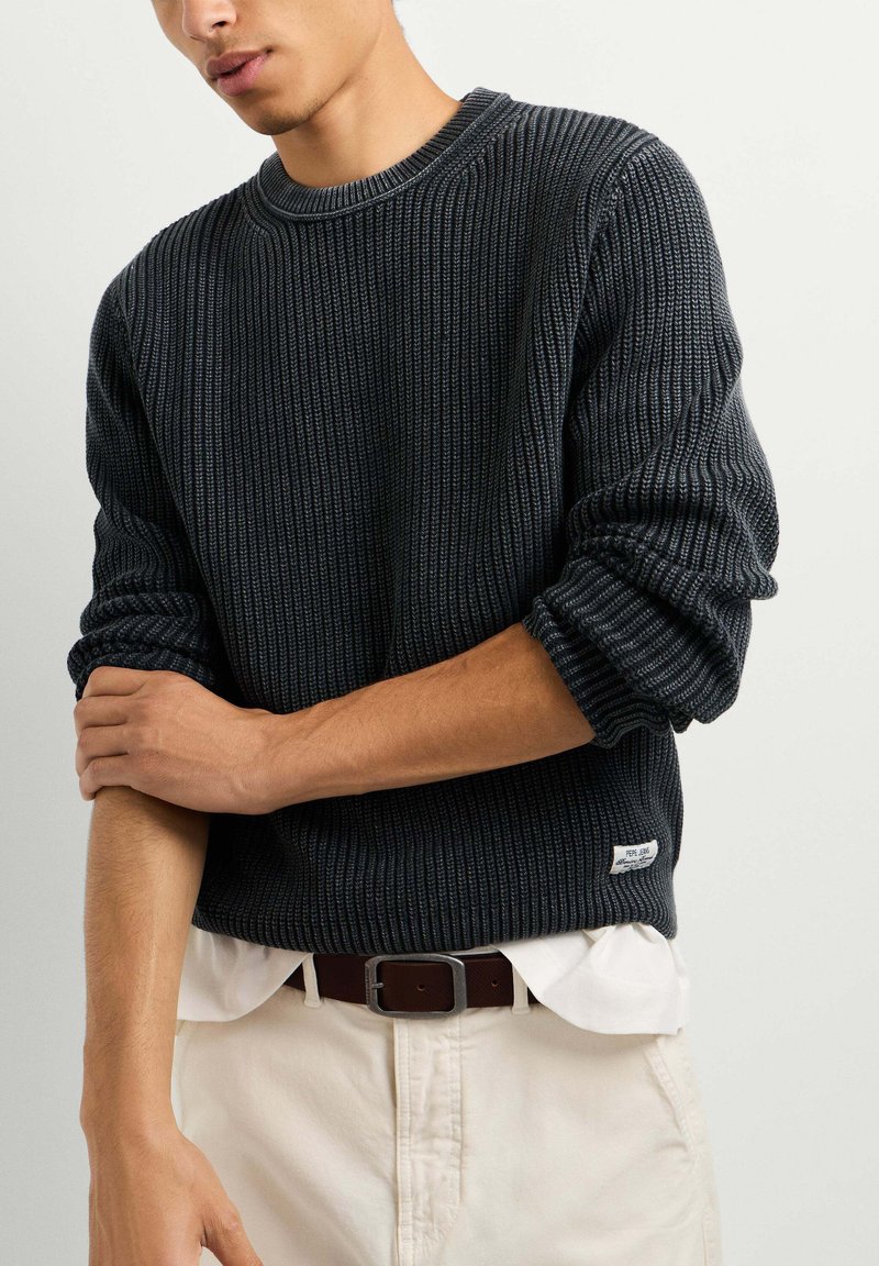 Personne portant un pull en maille côtelée foncée par-dessus une chemise blanche avec un pantalon beige et une ceinture sombre, ajustant une manche.