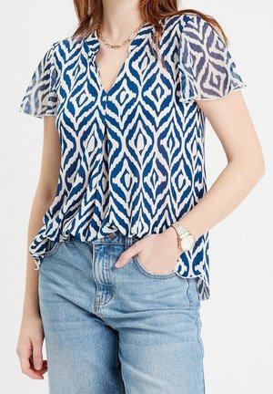Blouse à motifs bleus et blancs avec des manches courtes et transparentes, un décolleté en V, et un tissu léger et fluide ; associée à un jean en denim taille haute.
