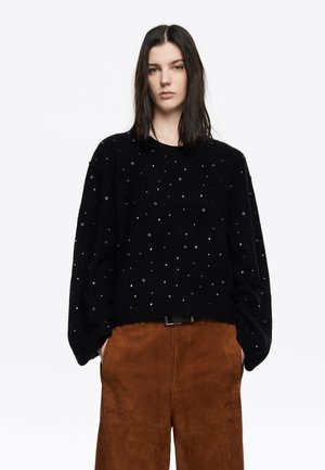 Suéter de punto negro con acentos metálicos plateados, formas redondas y triangulares. Ajuste relajado con hombros caídos, combinado con pantalones de ante marrones.