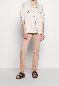 Vaaleanbeige farkkutakki, jossa on käännetyt hihat, yhdistettynä vaalepinkkeihin housuihin ja ruskeisiin sandaaleihin. Takissa on kaksi rintataskua ja napitus.