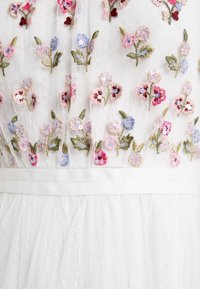 Valkoinen tüllimekko, jossa on kukkakirjailu vaaleanpunaisena, sinisenä ja vihreänä sekä paljetteja. Sisältää istuvan yläosan ja pehmeän, laskeutuvan hameen.