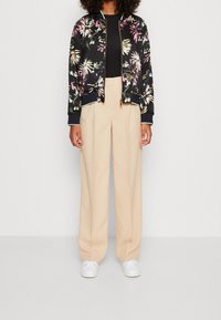 Bloemen bomberjack in zwart met veelkleurige palmprints, gecombineerd met lichtbeige wijde broeken en witte sneakers.