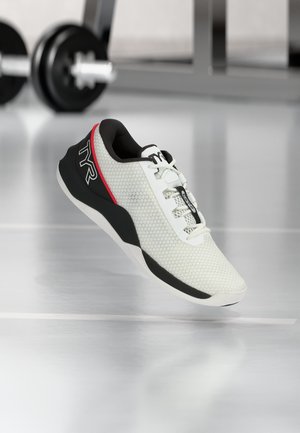 White and black athletic shoe with red accents and TYR logo, hovering above a reflective gym floor with blurred weights in the background.