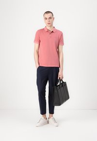 Korte mouwen polo in koraal, slim fit. Marineblauwe broek, tapered cut. Zwarte leren tote bag met dubbele handgrepen. Witte sneakers.
