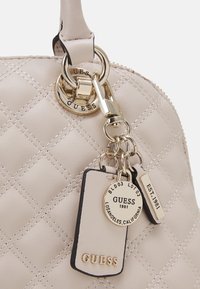 Beige quilted handbag with gold hardware, featuring a circular keychain and two tags, one circular and one rectangular with embossed text.