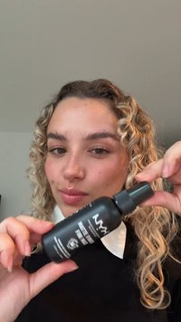 Black spray bottle with a pump top, labeled "NYX Matte Finish," held by a person with curly hair, featuring a matte finish and white text.