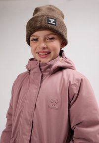 Roze waterdichte jas met een hoge kraag en geborduurd logo, gedragen met een bruine geribbelde beanie met een zwart label.