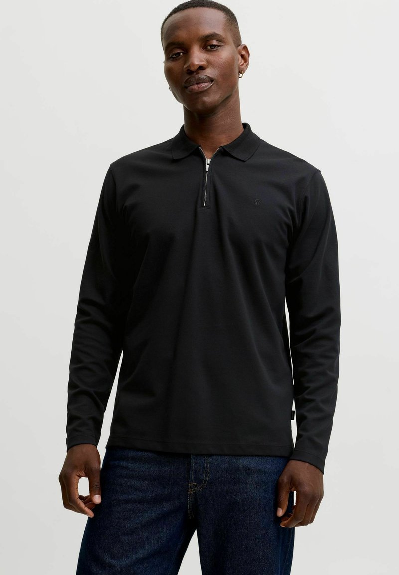 Homme portant un polo noir à manches longues avec une courte fermeture éclair et un jean bleu foncé, debout devant un fond clair uni.